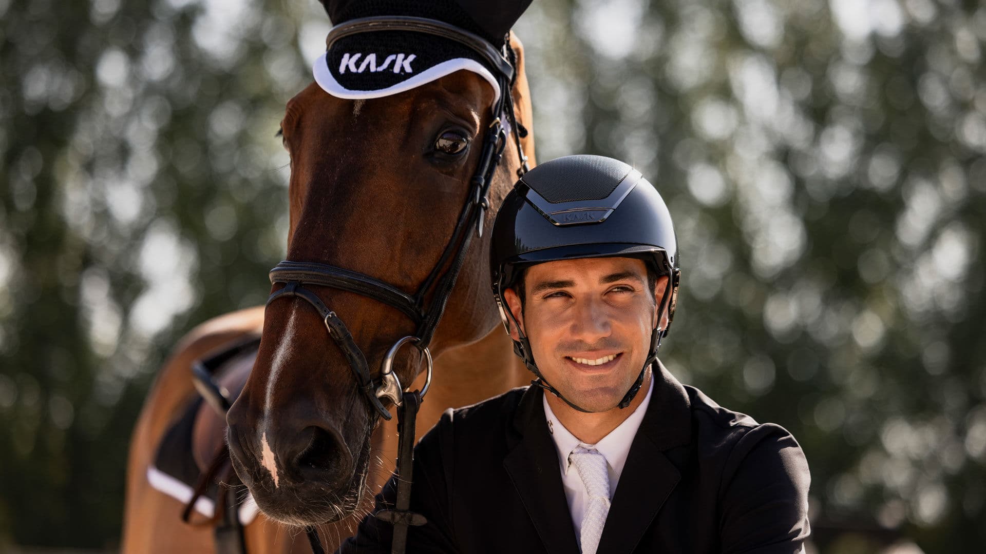 A rider in a black riding helmet smiles beside a chestnut horse.